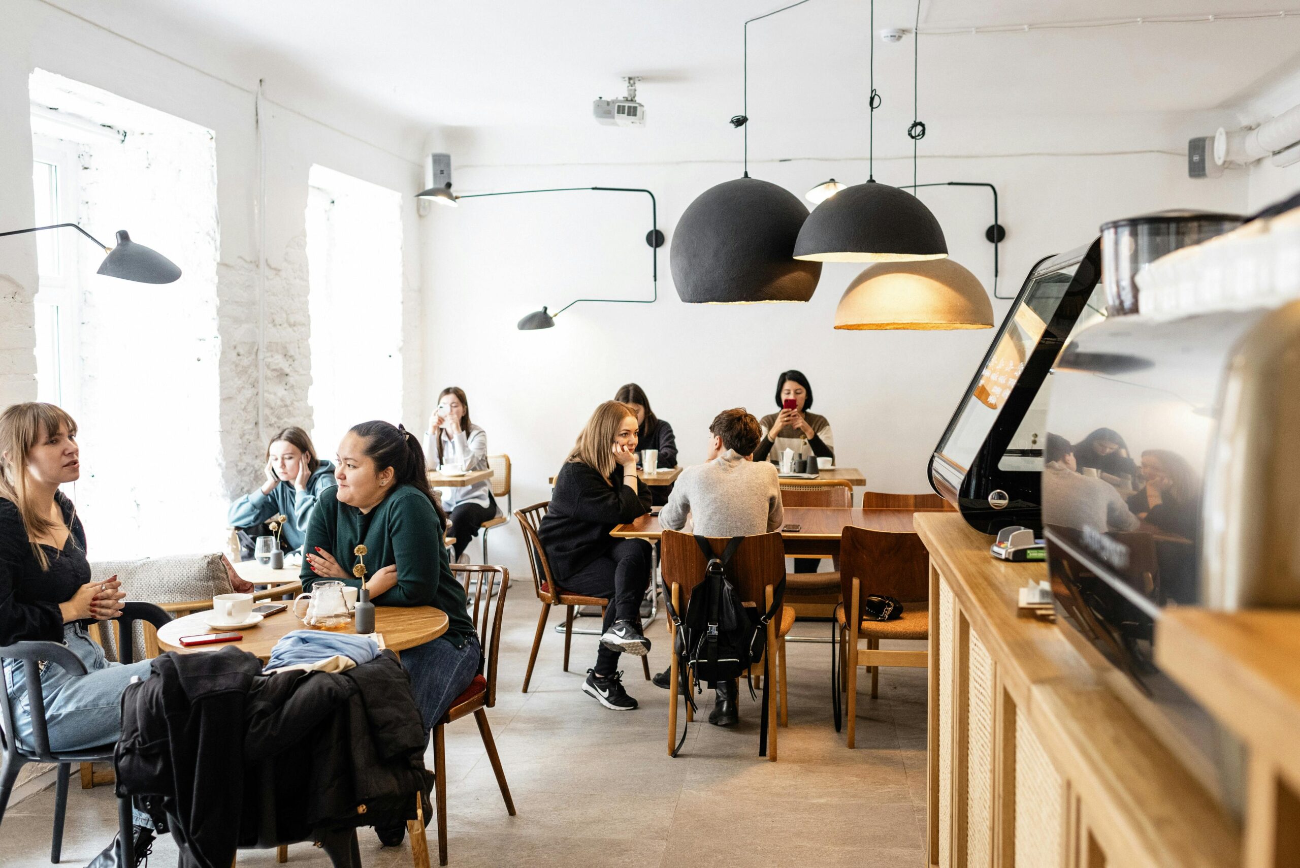 Group of multiracial friends in casual wear sitting at wooden tables in cozy cafeteria near cash box and big lamps while conversing